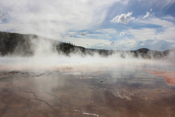 A vibrant hot spring is releasing steam into the air, surrounded by colorful mineral deposits. The clear blue sky and distant hills create a stunning backdrop in Yellowstone National Park.