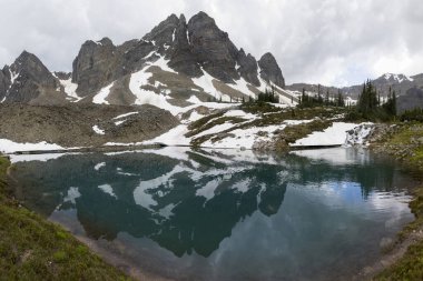 Sakin bir alp gölü engebeli bir dağ sırasının engebeli tepelerini yansıtır, seyrek yeşillik ve artık karla çevrilidir. Bulutlar dramatik bir gökyüzü yaratır.