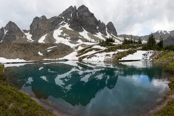 Sakin bir alp gölü engebeli bir dağ sırasının engebeli tepelerini yansıtır, seyrek yeşillik ve artık karla çevrilidir. Bulutlar dramatik bir gökyüzü yaratır.