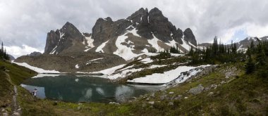 Sakin bir göl, bulutlu bir gökyüzünün altında yükselen dağları ve kar parçalarını yansıtır. Bereketli yeşillik suyu sarar, yürüyüş ve rahatlama için mükemmel bir doğal ortam yaratır..