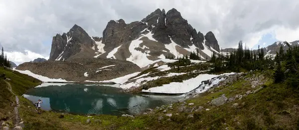 Sakin bir göl, bulutlu bir gökyüzünün altında yükselen dağları ve kar parçalarını yansıtır. Bereketli yeşillik suyu sarar, yürüyüş ve rahatlama için mükemmel bir doğal ortam yaratır..
