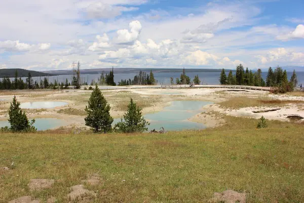 Yellowstone Ulusal Parkı 'nda geniş bir alanda yemyeşil jeotermal havuzlar ve bulutlarla dağılmış parlak mavi gökyüzünün altındaki uzak dağlar sergileniyor..