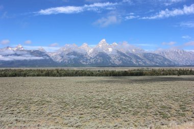 Wyoming 'de geniş bir çalı ovasıyla çevrili yüksek dağ zirveleri ufuk çizgisine hakim. Manzara parlak güneş ışığıyla aydınlanır, doğal güzellikleri sergiler..