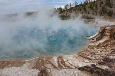Açık mavi sular kayalık bir kıyı şeridiyle çevrilidir ve Yellowstone Ulusal Parkı 'ndaki bir jeotermal kaplıcadan havaya buhar yükselir. Bu nefes kesici manzara doğanın güzelliğini gösteriyor..