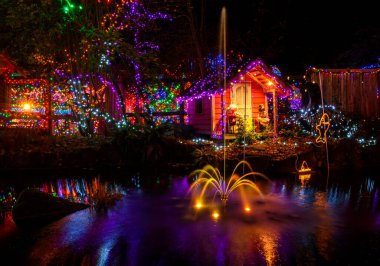 Büyüleyici bir kulübe, sakin bir gölete yansıyan canlı tatil ışıklarıyla süslenirken, bir çeşme büyüleyici akşam atmosferinde büyüleyici bir su özelliği yaratır..