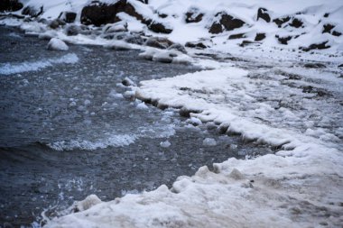 Dalgalar donmuş bir gölün karlı kıyılarına çarpar. Suyun kenarında buz oluşumları sakin ve serin bir ortamda sert kış manzarasını vurgular..
