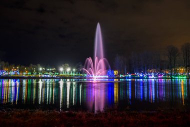 Canlı bir çeşme, sakin bir gölün kıyıları boyunca renkli ışıklarla çevrili pembe bir su fışkırtarak şenlikli bir atmosfer yaratır..