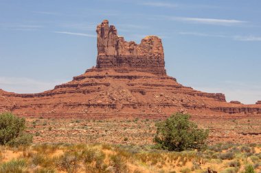 Utah, Monument Vadisi 'ndeki açık mavi gökyüzüne çarpan dramatik kırmızı kaya oluşumları. Güneş engebeli arazide gölgeler bırakır, doğanın güzelliğini gözler önüne serer.