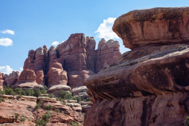 Canlı kırmızı kaya oluşumları açık mavi gökyüzünün altında kumtaşı katmanlarını gösteriyor. Bereketli yeşillik kayalık araziyi gözetliyor, gün ortasında engebeli doğal bir manzaranın özünü yakalıyor..