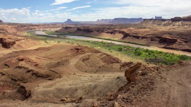 Colorado nehri, kırmızı kaya oluşumları ve yeşil bitki örtüsüyle geniş bir kanyon arazisi boyunca akar. Parlak mavi gökyüzü, doğal güzelliğe çarpıcı bir zemin sunar..