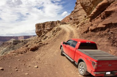 Kırmızı bir kamyonet Grand Canyon 'da engebeli bir toprak yolda ilerliyor. Dağınık bulutlarla dolu parlak bir gökyüzü altında nefes kesen kayalık manzarayı gösteriyor..