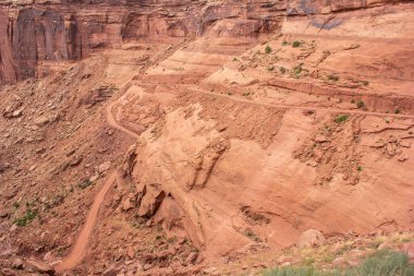 Moab, Utah 'taki görkemli kanyon manzaralarıyla çevrili sarmal bir toprak yolun dik kırmızı kaya oluşumları şeklinde oyulduğu görülebilir. Canlı renkler bölgenin eşsiz jeolojisini vurguluyor..