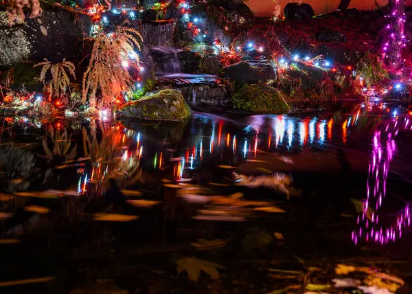 Huzurlu bir bahçe sahnesinde canlı ışıklar küçük bir şelaleyi aydınlatıyor. Su rengarenk parlaklığı yansıtır, gecenin sakin atmosferini güçlendirir..