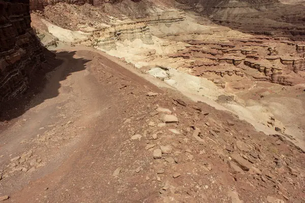 Dolambaçlı bir toprak yol, parlak güneş ışığı altında renkli kaya oluşumları gösteren geniş bir kanyon boyunca uzanır. Sahne, doğal manzaranın saf güzelliğini yakalar..