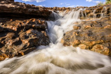 Kristal berraklığında su, sakin bir arazide engebeli kayaların üzerinden zarif bir şekilde akar. Güneş ışığı sahneyi aydınlatır, doğada barışçıl bir atmosfer yaratır..