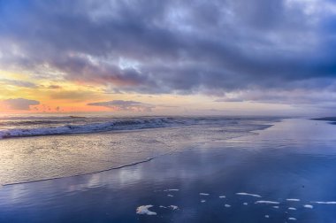 Renkli günbatımı gökyüzünü turuncu, pembe ve mor renkleriyle boyar. Okyanus dalgaları bulutlu bir gökyüzünün altında sakin bir kıyı şeridine nazikçe uzanır..