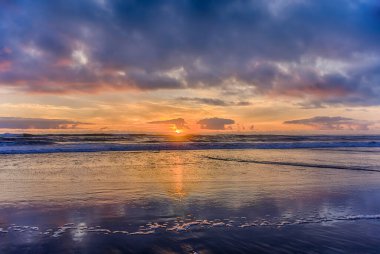 Güneş ufukta batarken dalgalar yavaşça kıyıya vurur, gökyüzüne sıcak renkler saçar ve kumsalın ıslak kumlarına yansıtır..