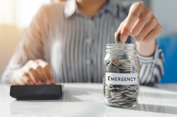 Hand of Young women putting Coins in a glass jar on the table. Saving for emergency concept.