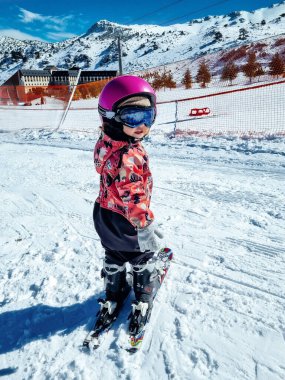 little child, girl 2 years old in pink ski suit, standing on skis, helmet and goggles on the descent from the mountain