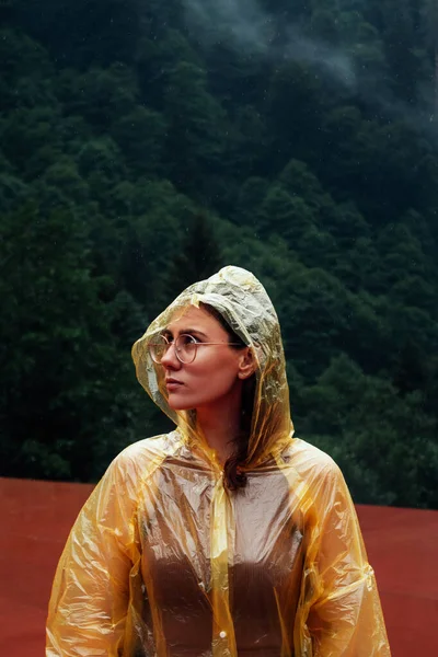 a girl in a yellow raincoat stands in the rain on the background of a dense forest of black pines, alone