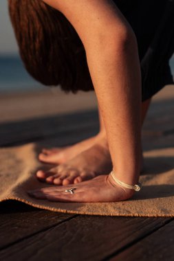 Denizin arka planındaki kız klasik yoga yapıyor. Diz çöküp, öne eğiliyor. Bileklik takıyor.