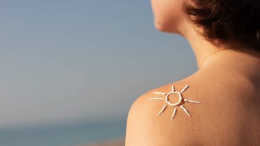 Güneş kremi, yazlık cilt bakımı. Kız sahilde denizin arka planında oturuyor. Omzunda kremle boyanmış bir güneş var.