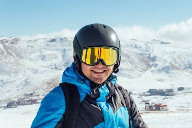 Kayakçı yamaçta duruyor. Mavi takım elbiseli adam, kayaktaki kask ve maske kayak yapmak içindir. Arkaplanda karla kaplı dağlar 