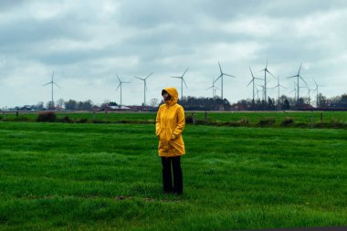 Sarı yağmurluklu bir kız, kara bulutlu yağmurlu havada yel değirmenlerinin arka planında duruyor.