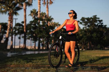 Açık hava takibi. sağlıklı yaşam tarzı konsepti. Bir kadın plajda gözlüklü bir spor bisikletiyle palmiye ağaçlarının arkasında dikiliyor.