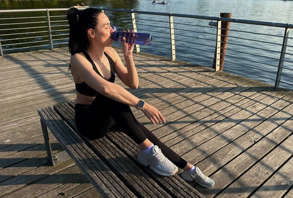 Drinking Water After Running. Portrait Of Beautiful Athletic Girl In Bright Colorful Sportswear Resting After Fitness Workout, Drink Water From Bottle On Blue Sky Background.