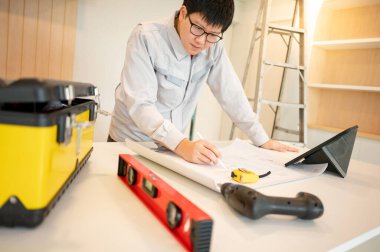 Ev tadilatı ya da tadilat. Yeni mutfak tezgahında mimari çizim ve inşaat aletleriyle çalışan Asyalı erkek iç işçisi veya mobilya toplayıcısı dijital tablet kullanıyor.