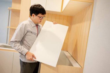 Ev tadilatı ya da ev dekorasyonu konsepti. Asyalı erkek mobilya derleyicisi ya da inşaat işçisi mutfak tezgahına dolap paneli yerleştiriyor. Yeni ev için mobilya kurulumu.