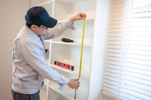 Home renovation or House remodeling concept. Asian male furniture assembler or Interior construction worker man using tape measure installing the new white modern cabinet.