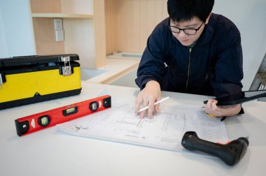 Ev tadilatı ya da tadilat. Yeni mutfak tezgahında mimari çizim ve inşaat aletleriyle çalışan Asyalı erkek iç işçisi veya mobilya toplayıcısı dijital tablet kullanıyor.