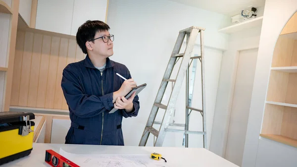 Home renovation or house remodeling. Asian male Interior worker using digital tablet working with construction tools on countertop of kitchen. Furniture assembler man installing cabinet for new house.
