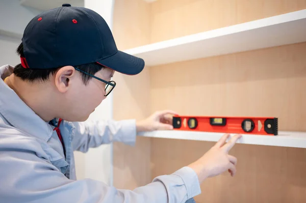Home renovation or House remodeling concept. Asian male furniture assembler or Interior construction worker man using spirit level tool installing white shelf in the new wooden cabinet.