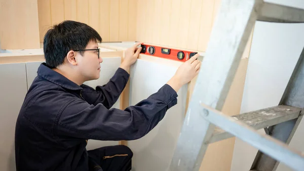 Home renovation or House remodeling concept. Asian male furniture assembler or Interior construction worker man using spirit level tool installing wooden counter and cabinet of new kitchen.
