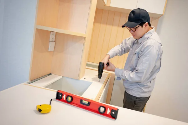 Home renovation or House remodeling concept. Asian male furniture assembler or Interior construction worker man using electric drill screwdriver installing wooden counter and cabinet of new kitchen.