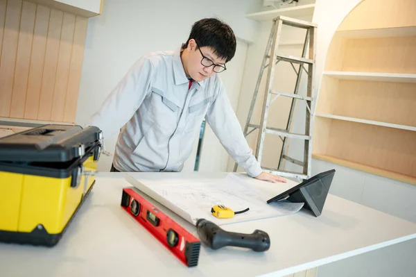 Home renovation or house remodeling. Asian male Interior worker or furniture assembler man using digital tablet working with architectural drawing and construction tools on countertop of new kitchen.