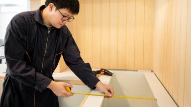 Ev tadilatı ya da ev dekorasyonu konsepti. Asyalı erkek mobilya derleyicisi ya da inşaat işçisi yeni mutfağın ahşap tezgah ve dolabını bant ölçüsüyle kuruyor..