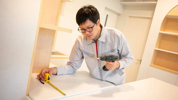 Home renovation or House remodeling. Asian male furniture assembler or Interior construction worker man using tape measure and electric screwdriver installing shelf, cabinet and kitchen counter.