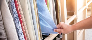 Kumaş kumaşından mavi kumaş parçası seçen stilist el. Döşeme örneği koleksiyonu vitrin rafında asılı duruyor. Ev iç tasarımı ve mobilya dekorasyonu için tekstil endüstrisi