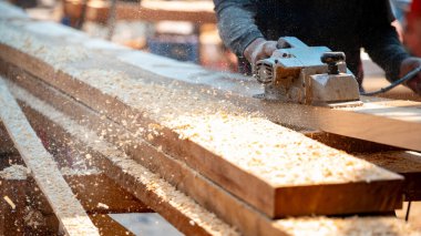 Erkek marangoz inşaat alanında tahta üzerinde elektrikli bir planör kullanıyor. Atölyesinde kereste planlayan ahşap işçisi etrafa talaş saçıyor. Kereste endüstrisi ve marangozluk çalışma aracı