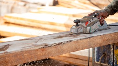 Erkek marangoz, atölyedeki tahta üzerinde elektrikli bir planör kullanıyor. İnşaat alanında kereste planlayan ahşap işçisi etrafa talaş saçıyor. Marangozluk aleti. Kereste endüstrisi konsepti