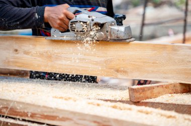 Erkek marangoz, atölyedeki tahta üzerinde elektrikli bir planör kullanıyor. İnşaat alanında kereste planlayan ahşap işçisi etrafa talaş saçıyor. Marangozluk aleti. Kereste endüstrisi konsepti