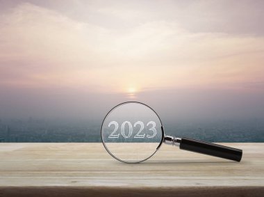 2023 yazı ve mali grafiği olan büyüteç modern şehir kulesinin üzerinde ahşap masa ve günbatımında gökdelen, klasik tarz, iş yeni yılı 2023 araştırma konsepti.