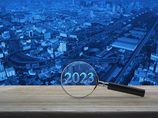 Magnifying glass with 2023 letter and financial graph on wooden table over modern office city tower, street, expressway, and skyscraper, Business happy new year 2023 research concept