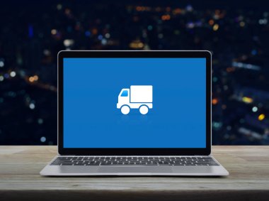Delivery truck flat icon on modern laptop computer screen on wooden table over blur colorful night light office city tower and skyscraper, Business transportation online service concept