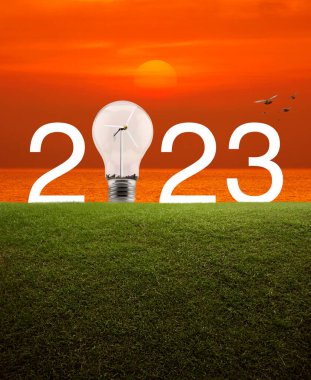 2023 beyaz yazı ve ampul içinde rüzgar türbini olan yeşil çimlerin üzerinde günbatımı gökyüzü ve denizin üzerinde kuşlarla birlikte. Mutlu yeni yıl 2023 ekolojik kapak kavramı.