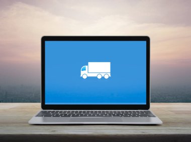 Delivery truck flat icon on modern laptop computer screen on wooden table over office city tower and skyscraper at sunset sky, vintage style, Business transportation online service concept